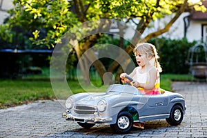 Little adorable toddler girl driving big vintage toy car and having fun with playing outdoors. Gorgeous happy healthy