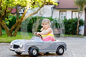 Little adorable toddler girl driving big vintage toy car and having fun with playing outdoors. Gorgeous happy healthy