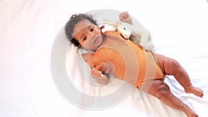 a little African-American baby girl in an orange bodysuit on the bed at home, a funny six-month-old black newborn baby