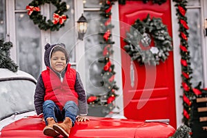 Little african american boy sitting on the hood of Christmas vintage car. Child fooling around
