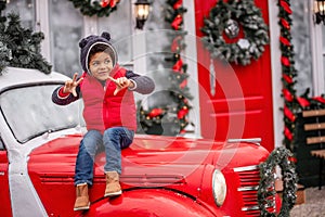 Little african american boy sitting on the hood of Christmas vintage car. Child fooling around