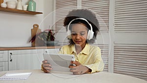 Little African American girl in big white headphones plays a game using a digital tablet. Rest during online learning