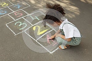 Little African American girl drawing hopscotch with chalk on asphalt outdoors. Happy childhood