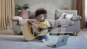 Little African American girl learning to play acoustic guitar while looking at laptop screen. Teenage girl sits on floor