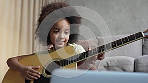 Little African American girl learning to play guitar while looking at laptop screen. Teenage girl sits on floor in hall