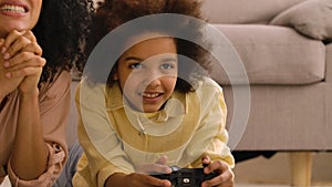 Little African American girl plays game on wireless joystick and rejoices victory. Mother and daughter lying on floor in