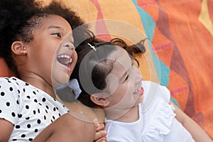 Little African and Caucasian kids girls smiling laughing and lying together on colorful mat on green grass