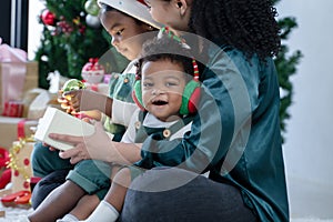 Little African child boy is sitting smiling in arms of Asian young mother with earmuffs enjoy celebrating Christmas at home