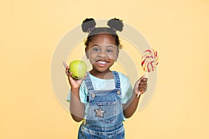 Little African child girl holds sweet lollipop and fresh green apple, smiling to camera on yellow background. Healthy
