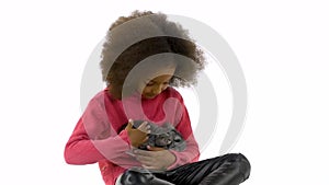 Little african curly girl is holding and stroking grey chinchilla at white background. Slow motion