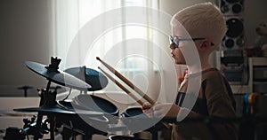 A little albino boy with white hair color in blue round glasses plays on an electronic drum set at home in his room