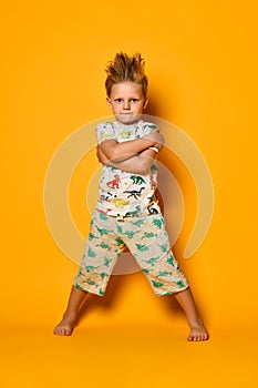 Angry blond boy preschooler in pajamas with dinosaurs having fun has joy in the studio over yellow background.