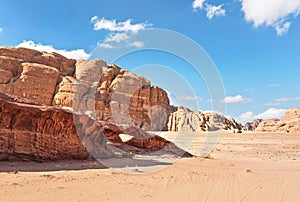 Little arc or small rock window formation in Wadi Rum desert, bright sun shines on red dust and rocks, blue sky above