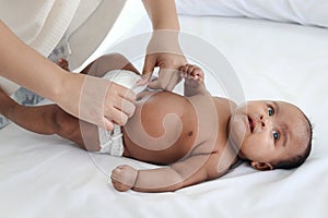 A little Asian African newborn baby girl lying on white bed with mother hands changing baby nappy, mom putting diaper on her sweet