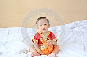 Little Asian baby boy in traditional Chinese dress with a piggy bank sitting on bed at home. Kid saving money concept