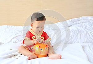 Little Asian baby boy in traditional Chinese dress putting some coins into a piggy bank sitting on bed at home. Kid saving money