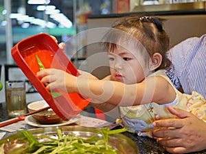 Little Asian baby girl, 3 years old, enjoys putting vegetables in to a hot pot - engaging baby into food preparation