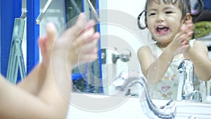 Little Asian baby girl enjoys opening a water tap, playing water, and washing her hands in front of a mirror by