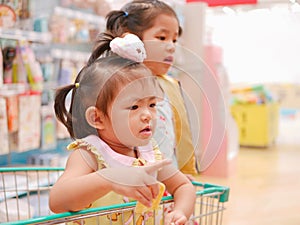 Little Asian baby girl having fun standing in a shopping cart with her sister seeing many products for sale