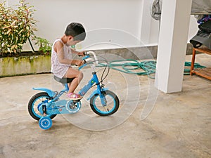 Little Asian baby learning to ride a bicycle with training wheels at home