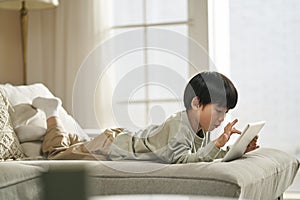 little asian boy lying on front on couch playing computer game using digital tablet