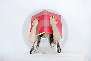 Little Asian child girl hiding face with open hardcover book sitting on bed with pillow