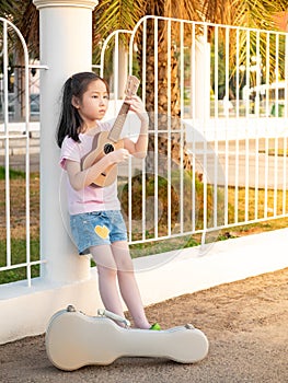 Little asian child girl play the ukulele, in the garden, leaning against the fence, practice to play