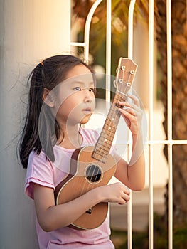 Little asian child girl play the ukulele, in the garden, leaning against the fence, practice to play