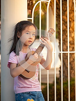 Little asian child girl play the ukulele, in the garden, leaning against the fence, practice to play, in sunset light