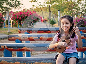 Little asian child girl play the ukulele, in the garden on the Steel pipem, sunset time