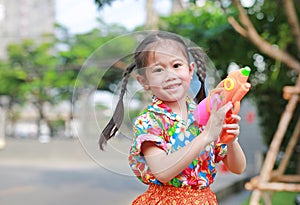 Little Asian child girl in traditional flower dress playing water gun at Songkran festival Thai new year - water festival