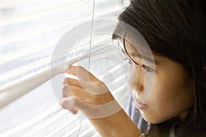 Little Asian girl cautiously peeking out window through blinds