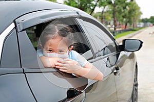 Little Asian girl child wears hygiene face mask looking through camera with pokes her head out of car window