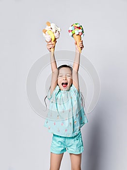 Little asian girl kid with eyes closed with happiness holds up high two sweet big ice-cream in waffles cones with tasty toppings