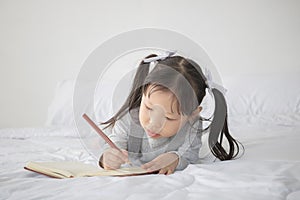 Little Asian girl lying on bed and writing alphabet on notebook