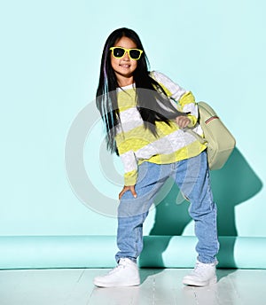 Little Asian girl model posing in the studio on a background of mint with a backpack on her shoulders. The girl wears a sweater,