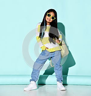 Little Asian girl model posing in the studio on a background of mint with a backpack on her shoulders. The girl wears a sweater,
