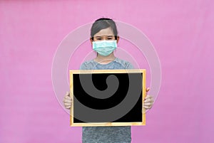 Little asian girl in a protective medical mask holding black board isolated on pink background.