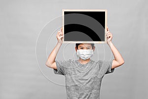 Little asian girl in a protective medical mask holding blackboard isolated on gray background.