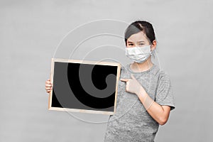 Little asian girl in a protective medical mask holding blackboard isolated on gray background.