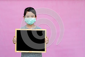 Little asian girl in a protective medical mask holding blackboard isolated on pink background.
