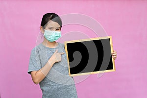 Little asian girl in a protective medical mask holding blackboard isolated on pink background.