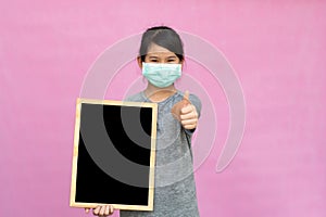 Little asian girl in a protective medical mask holding blackboard isolated on pink background.