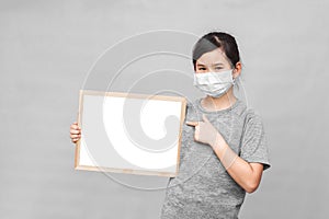 Little asian girl in a protective medical mask holding whiteboard isolated on gray background.