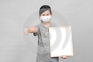 Little asian girl in a protective medical mask holding whiteboard isolated on gray background.