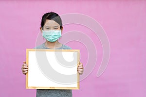 Little asian girl in a protective medical mask holding whiteboard isolated on pink background.