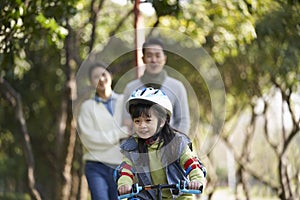 Little asian girl riding bike in city park with parents in background