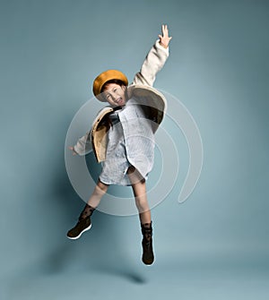 Little asian girl in shirt dress, double sided jacket, brown beret, boots. Laughing, jumping up on blue background. Full length