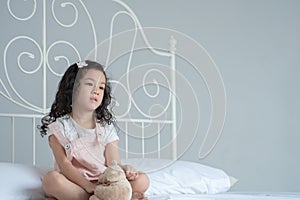 Little Asian girl sitting on bed with a teddy bear thinking and looking out of her bedroom