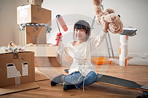Little asian girl smiling, sitting on boosted board, holding teddy bear and paint roller, with hands in the air, surrounded with photo